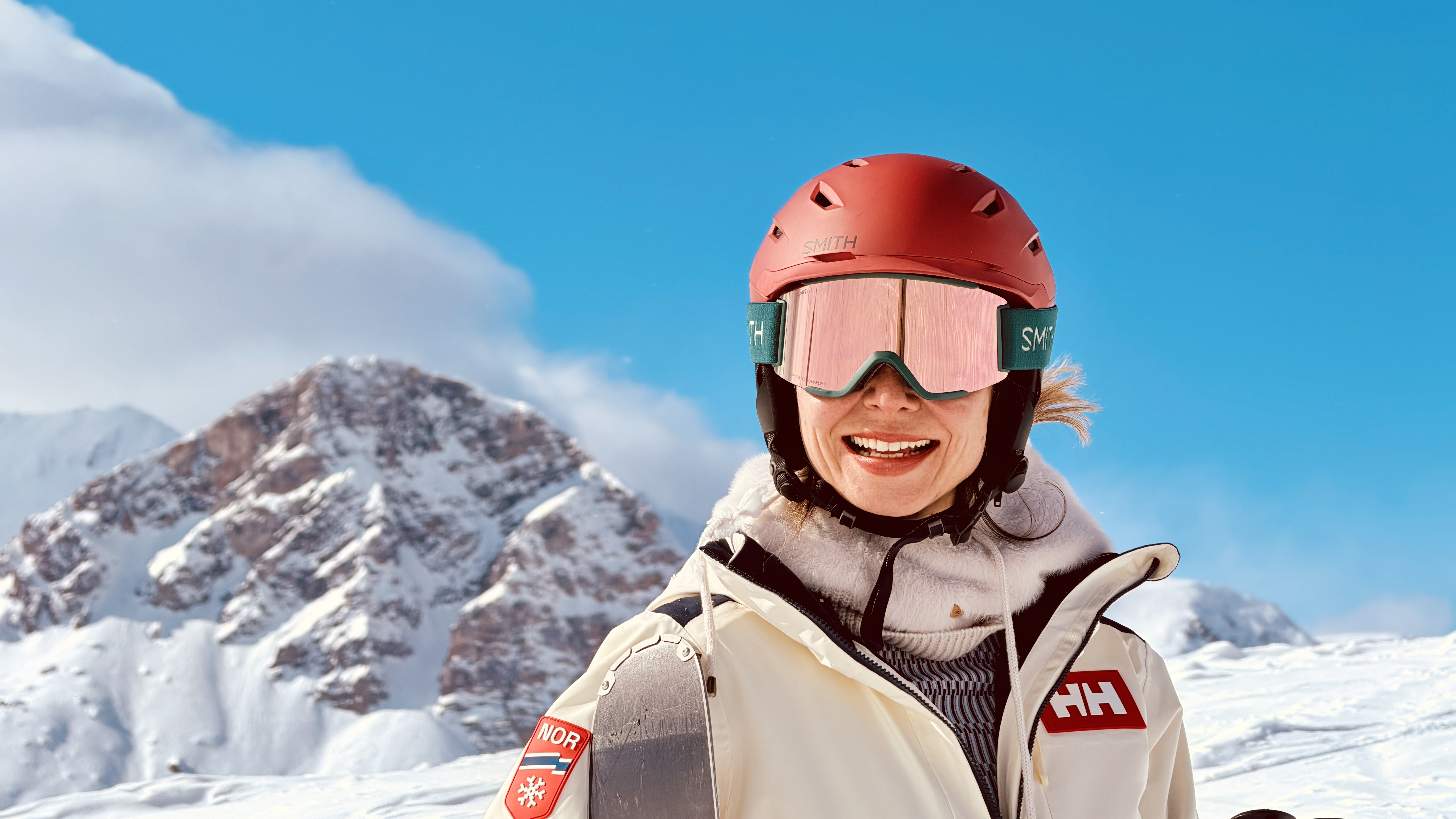Smiling skier in a red helmet and pink goggles in front of a snowy peak