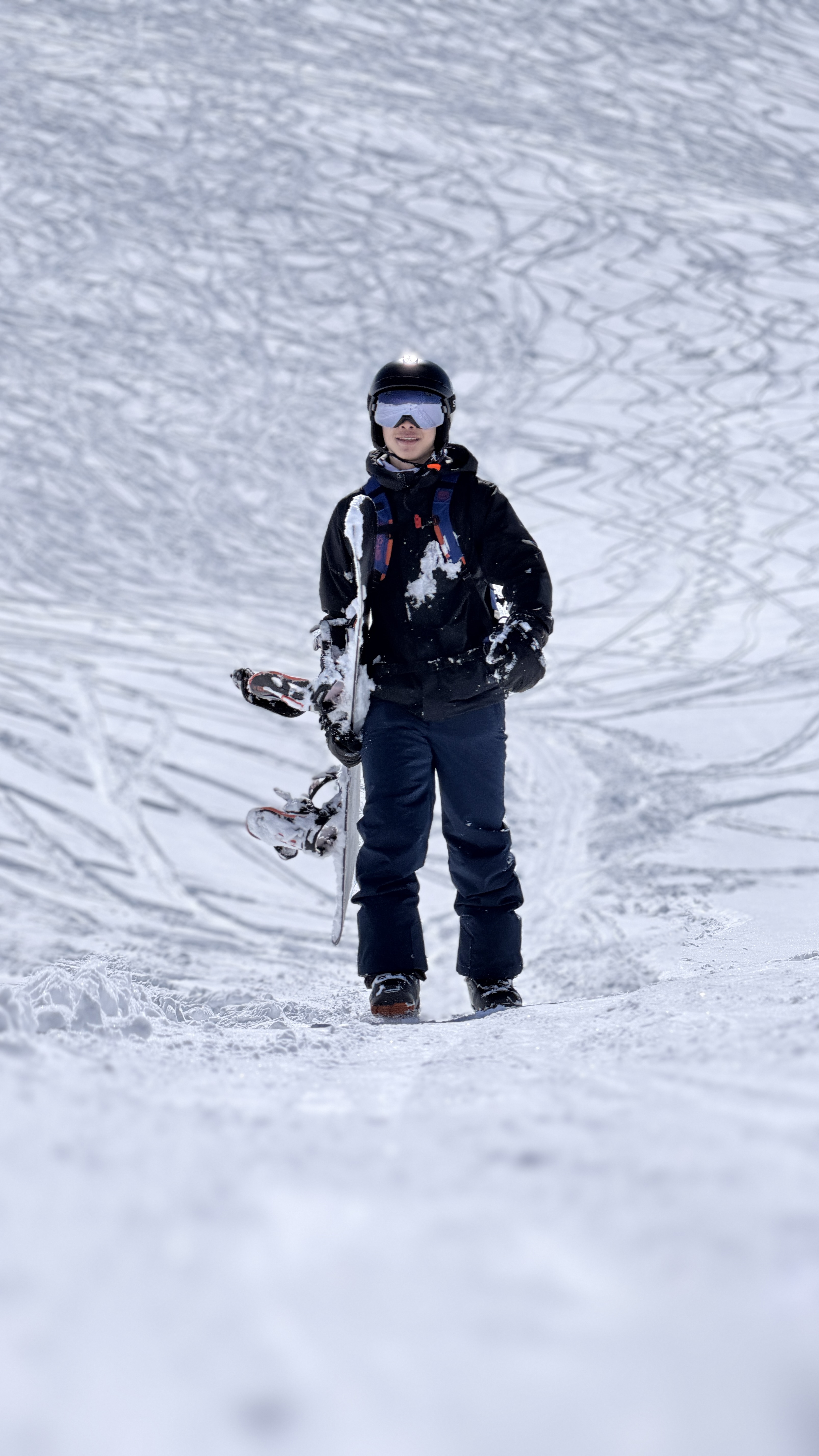 Snowboarder hiking up a slope covered in ski tracks, board in hand
